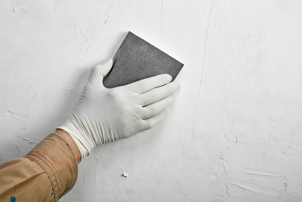 Een persoon schuurt een gestucte muur met een schuurpapier.